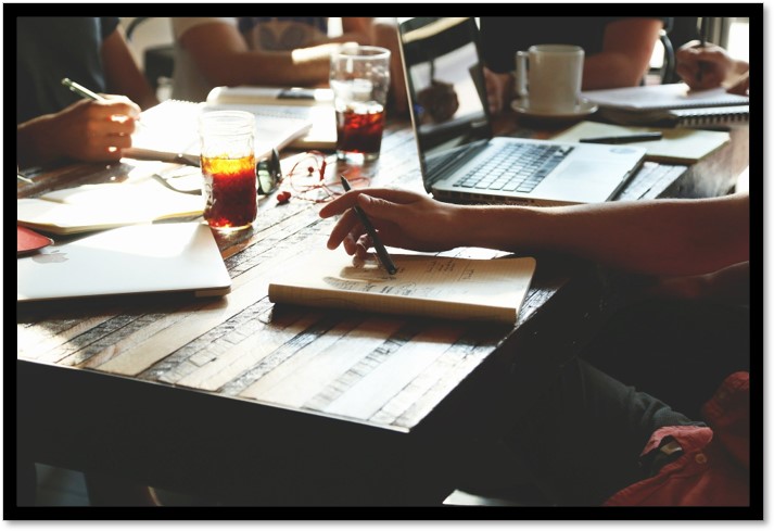 close up of table filled with coffee, papers, computers and hands with pencils visible