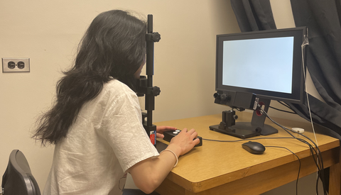 A student sitting at the desk in from of the monitor and an eye tracking camera holding a button box in her hands