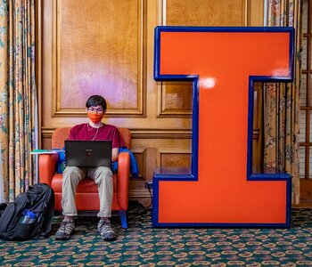 Setting in the Illini Union, on left a student sitting in an arm chair working on their laptop, on right a large orange letter "I"