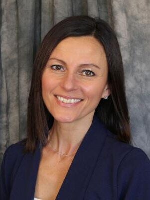 A headshot of Nikki Mercer in a blue blazer smiling in front of a grey fabric background