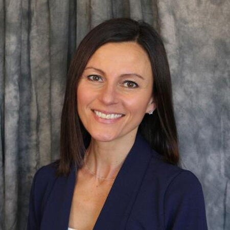 A headshot of Nikki Mercer in a blue blazer smiling in front of a grey fabric background