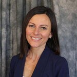 A headshot of Nikki Mercer in a blue blazer smiling in front of a grey fabric background